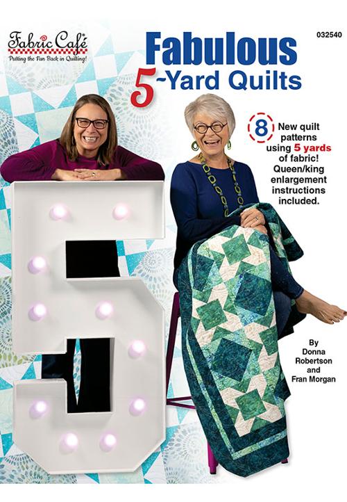 Two women with a quilt and a large letter 'F' at a quilt show, featuring Fabrics Cafe's 'Fabulous 5-Yard Quilts' book.