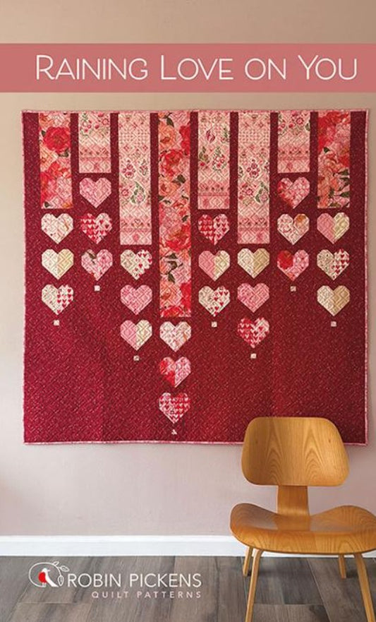 Quilt with heart patterns hanging on a wall above a wooden chair, with 'Raining Love On You' text and Robin Pickens branding.
